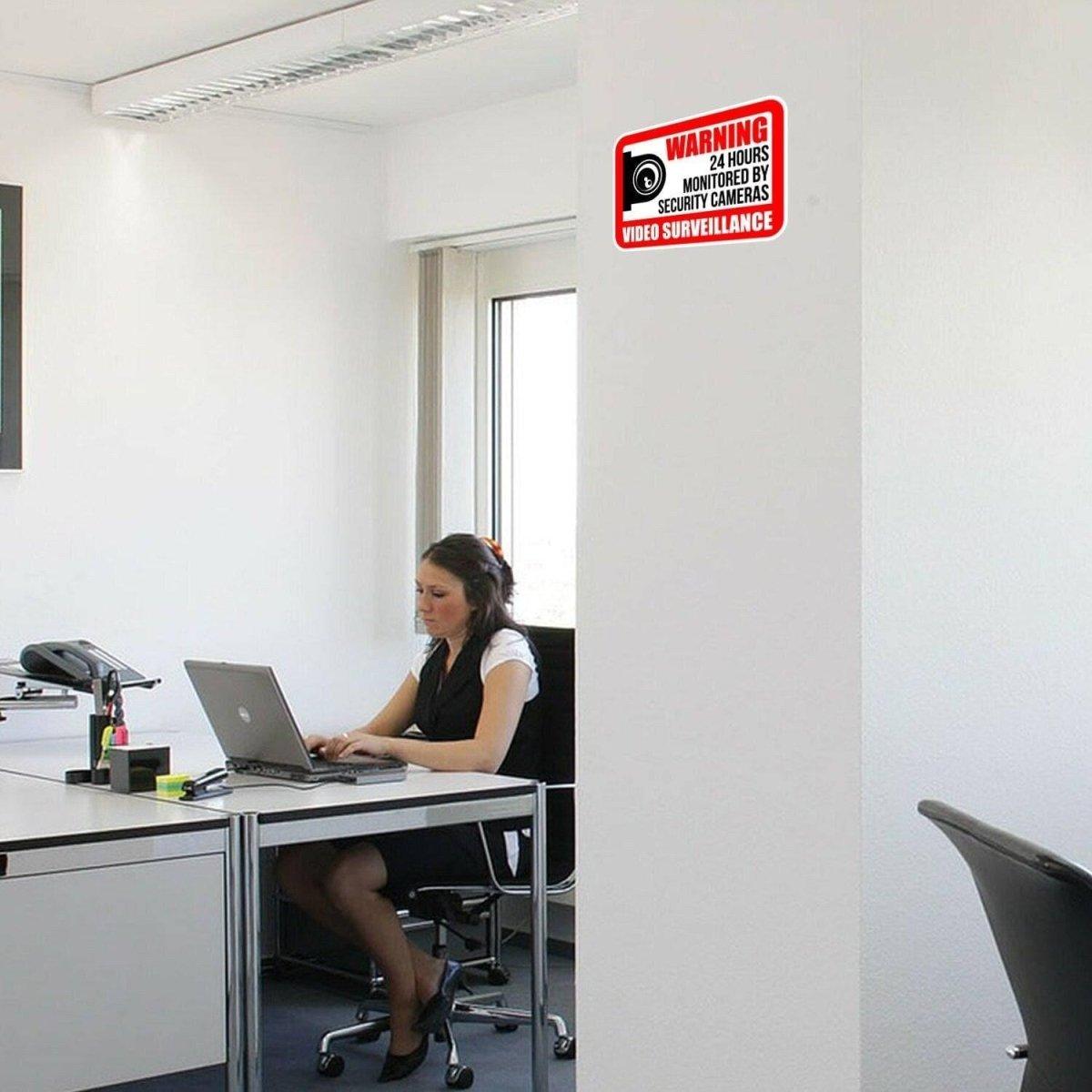 Office setting with woman at desk and warning sign about video surveillance on the wall.