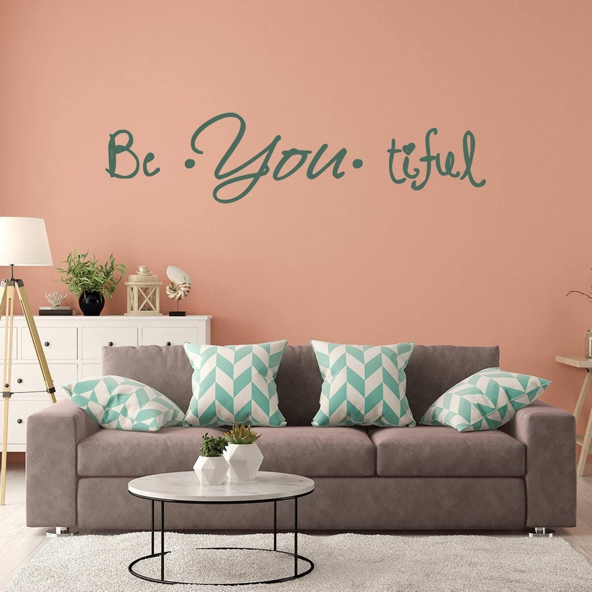 Motivational wall decal 'Be You tiful' on a peach wall above a stylish gray couch with blue-green patterned pillows.
