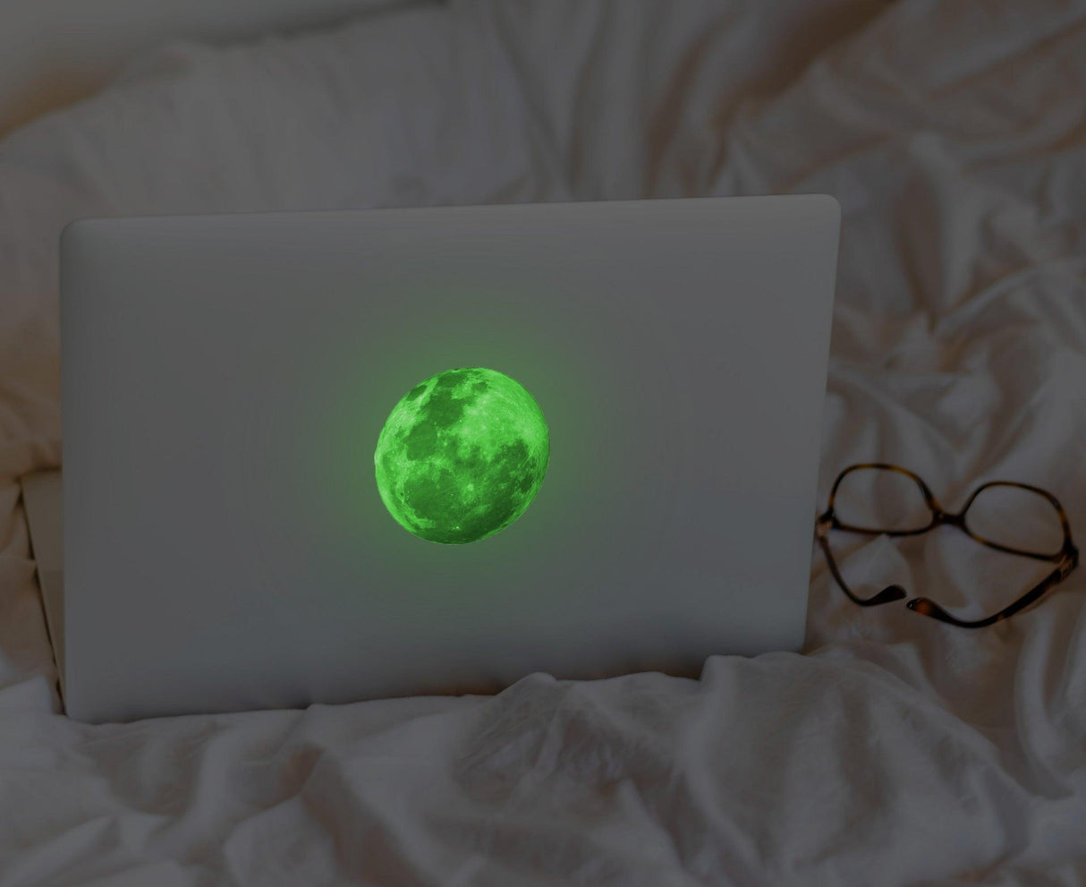 Glow in the dark moon decal on a laptop resting on a bed, surrounded by cozy bedding and glasses.