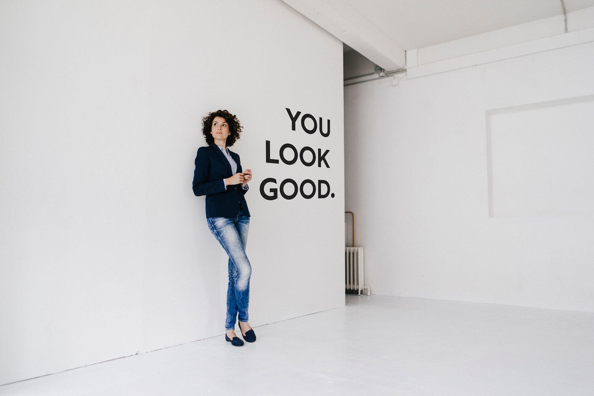 Woman in a blazer standing beside a 'You Look Good' vinyl wall sticker in a minimalist room.