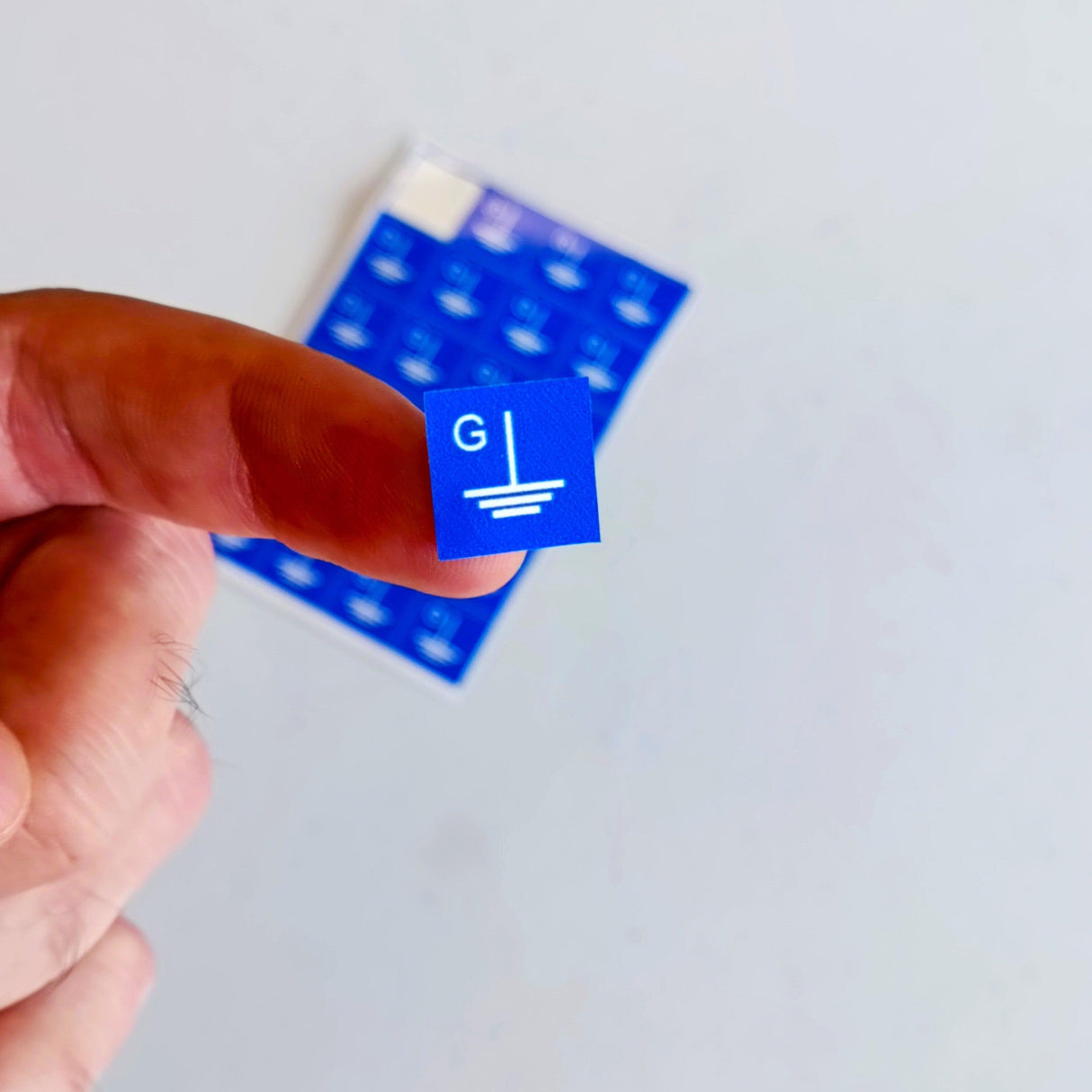 Blue grounding symbol sticker held between fingers, showcasing electrical component labeling for safety.