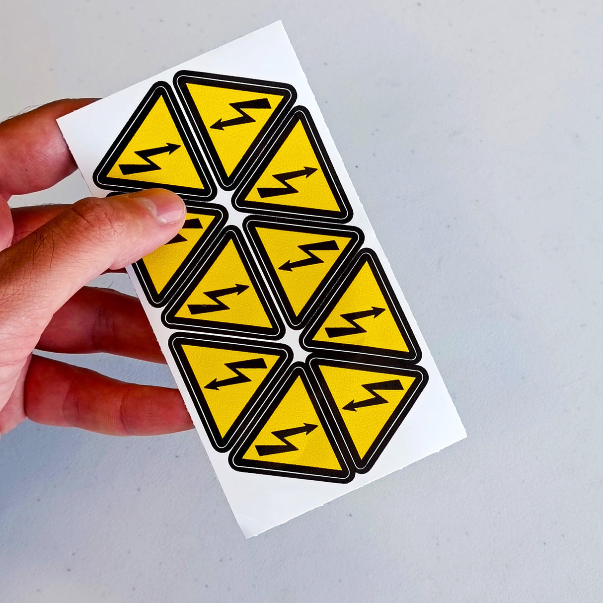 Set of 10 high voltage warning stickers in hand, featuring yellow triangle decals with lightning bolt symbol.