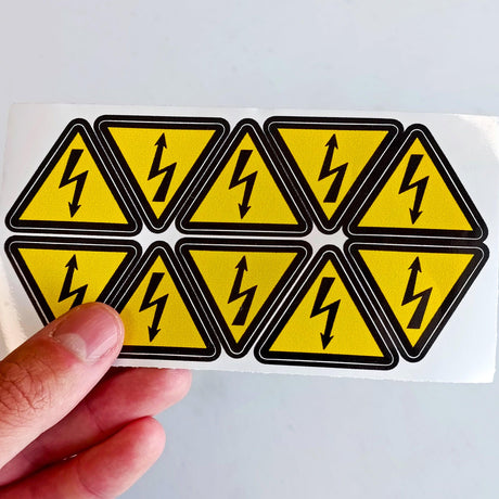 High Voltage Electrical Shock Hazard Stickers - Pack of 10 yellow triangle warning vinyl decals held in hand.