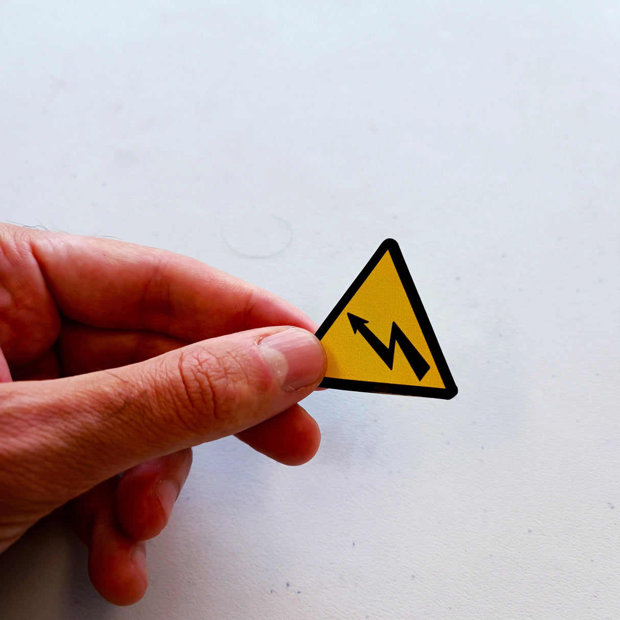 Hand holding a yellow triangle warning sticker for high voltage electrical shock hazards.
