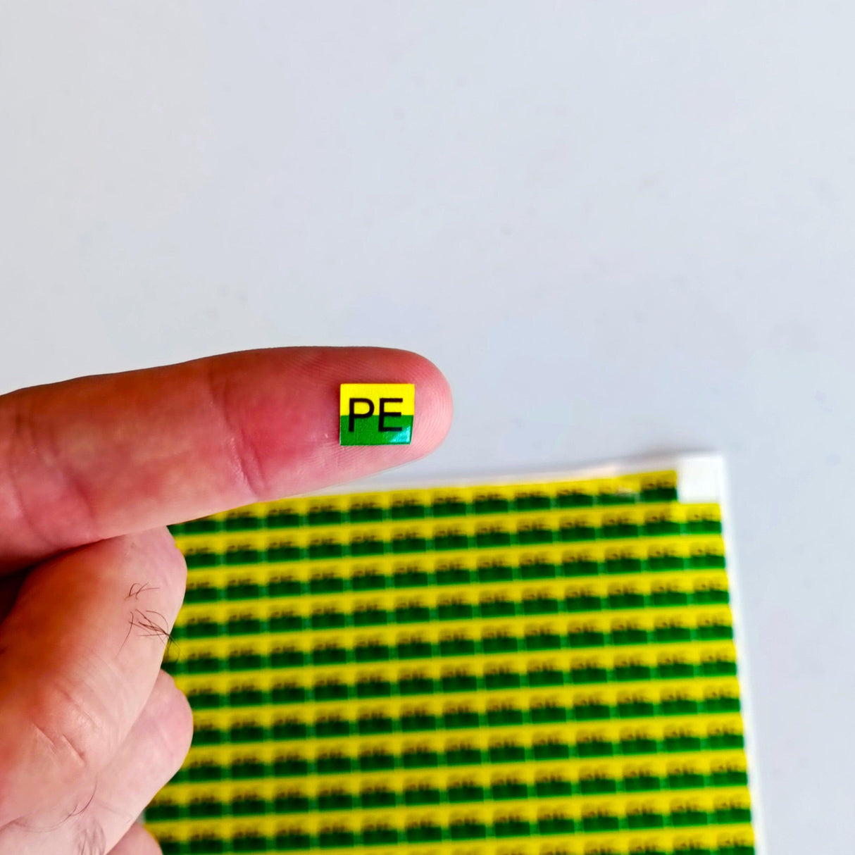 Close-up of a protective earth (PE) sticker held on a fingertip, with vibrant yellow and green backing.
