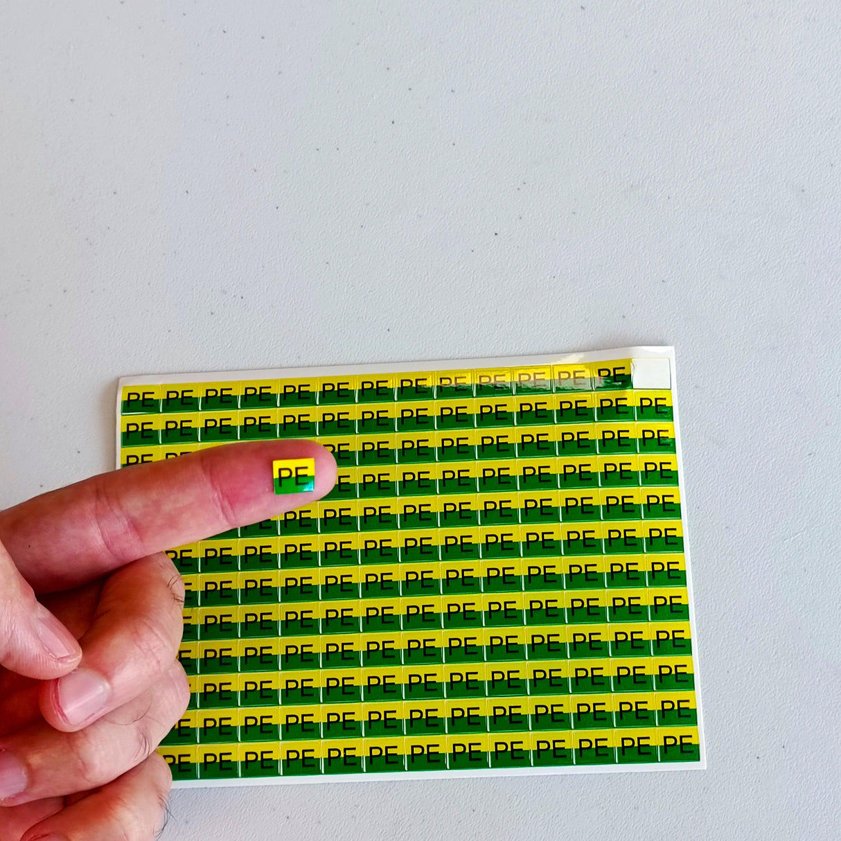 Hand holding a yellow and green PE electrical labeling sticker over a sheet of PE marking stickers.