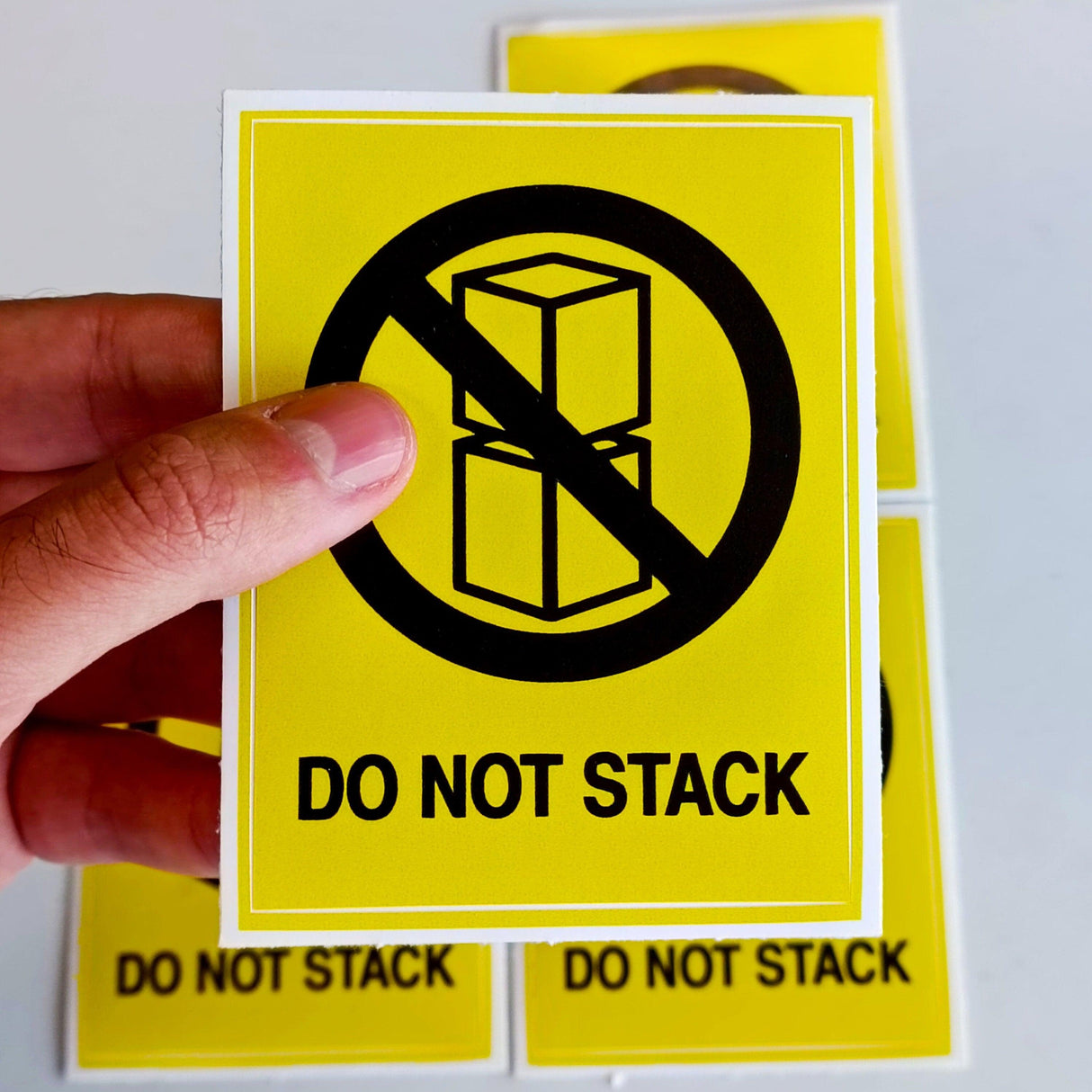 Bright yellow 'Do Not Stack' warning label sticker being held, designed for warehouse and shipping safety.