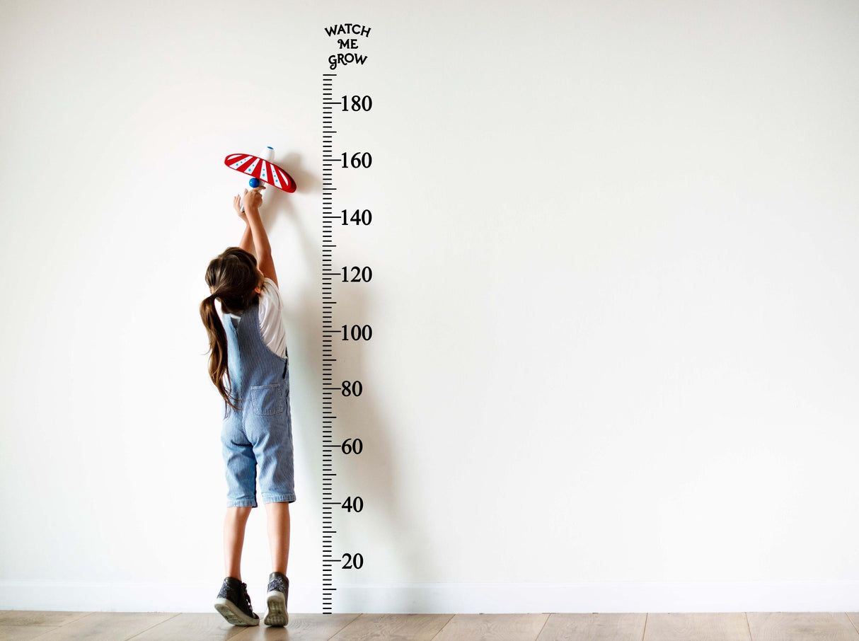 Child reaching for a vinyl growth chart ruler wall decal measuring centimeters in a bright room.