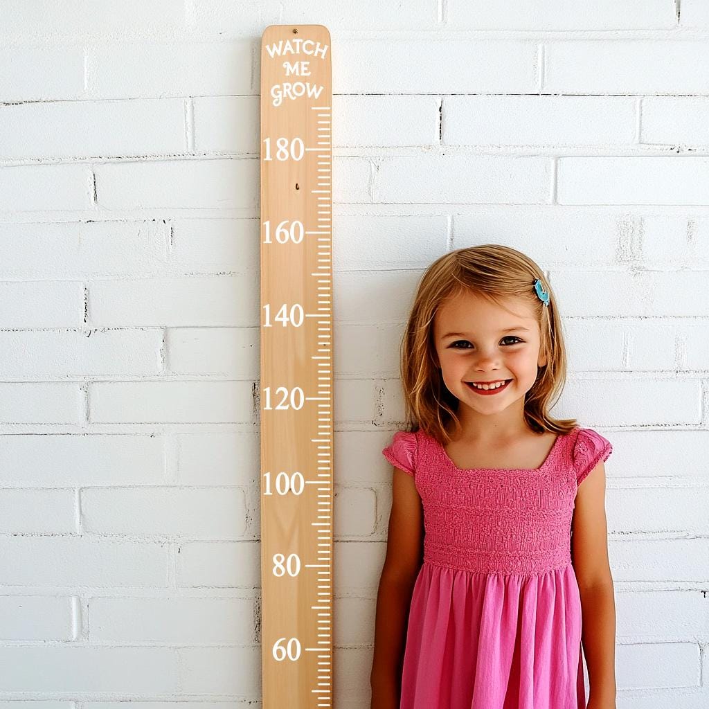 A smiling girl in a pink dress stands beside a wooden growth chart decal on a white wall.