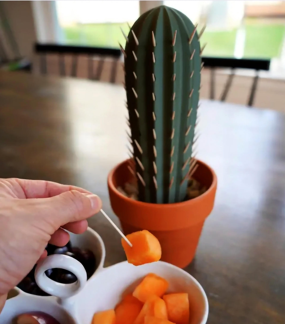 Cactus toothpick holder on a table with toothpick in hand and fruit pieces for a fun party accessory.