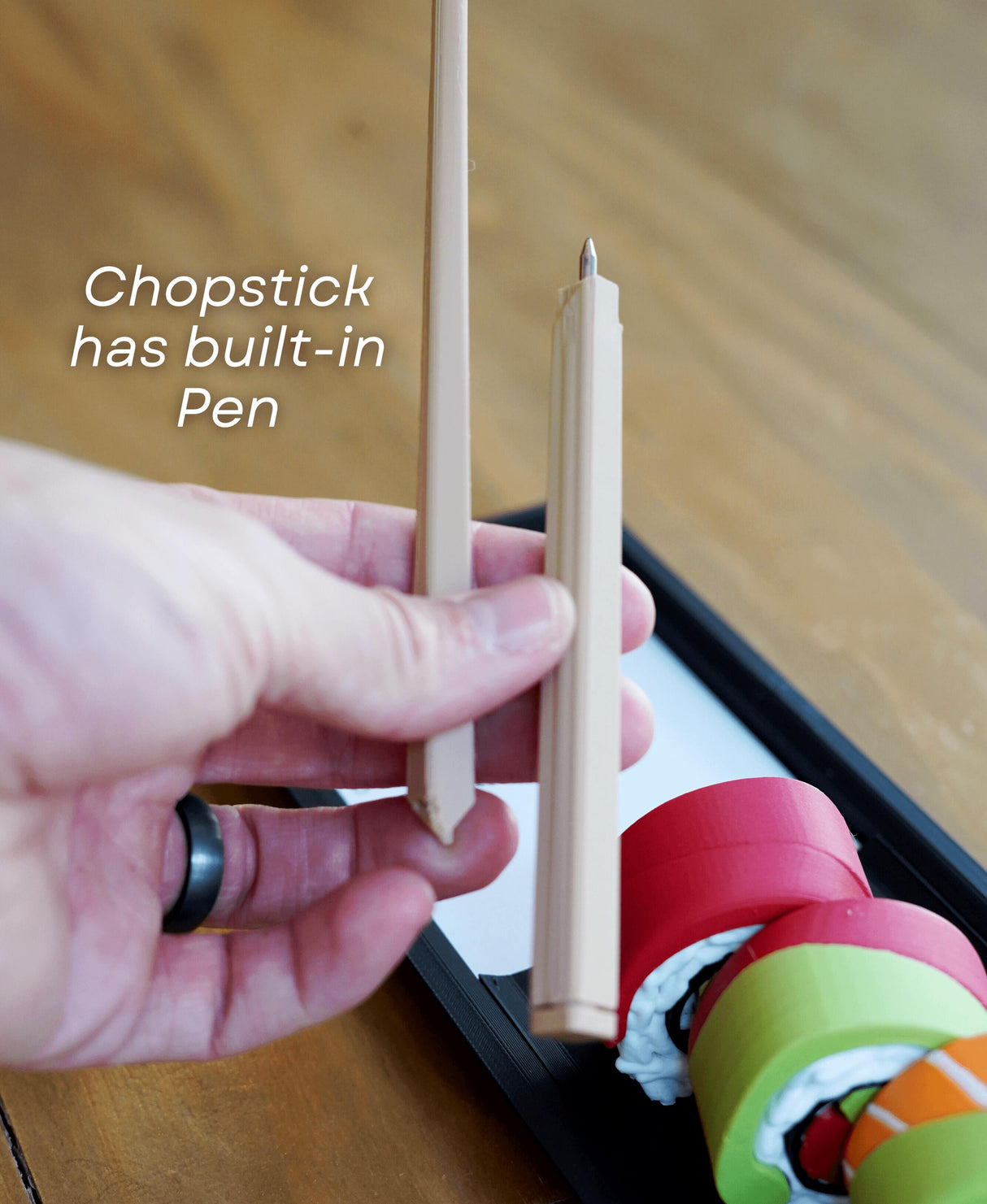 Hand holding chopstick that doubles as a pen alongside colorful sushi containers in a tray.