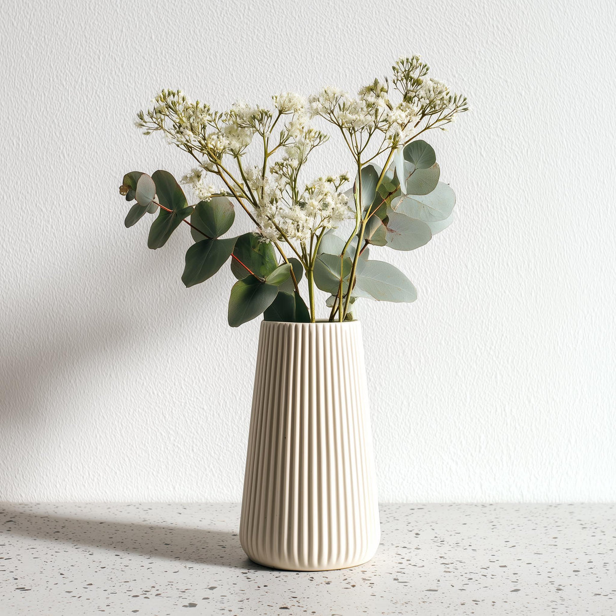 A tall, cream-colored 3D printed origami vase with vertical ribbed lines, holding white dried flowers and eucalyptus stems, placed on a speckled countertop.