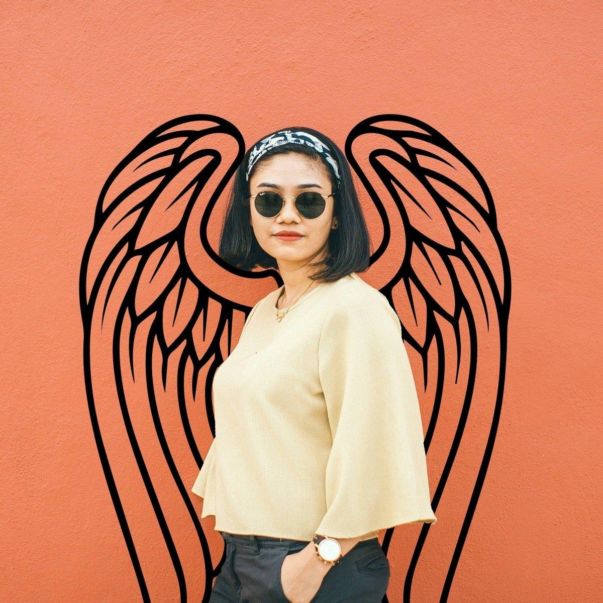 Woman in sunglasses standing against an orange wall with angel wings decoration in the background.