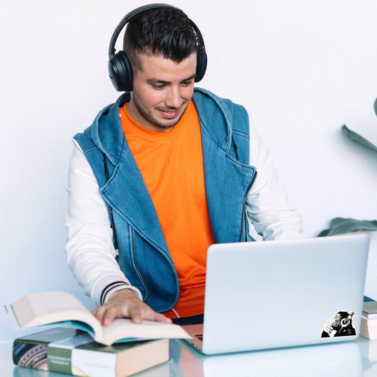 Young man with headphones using a laptop featuring a glowing monkey vinyl decal, surrounded by books.