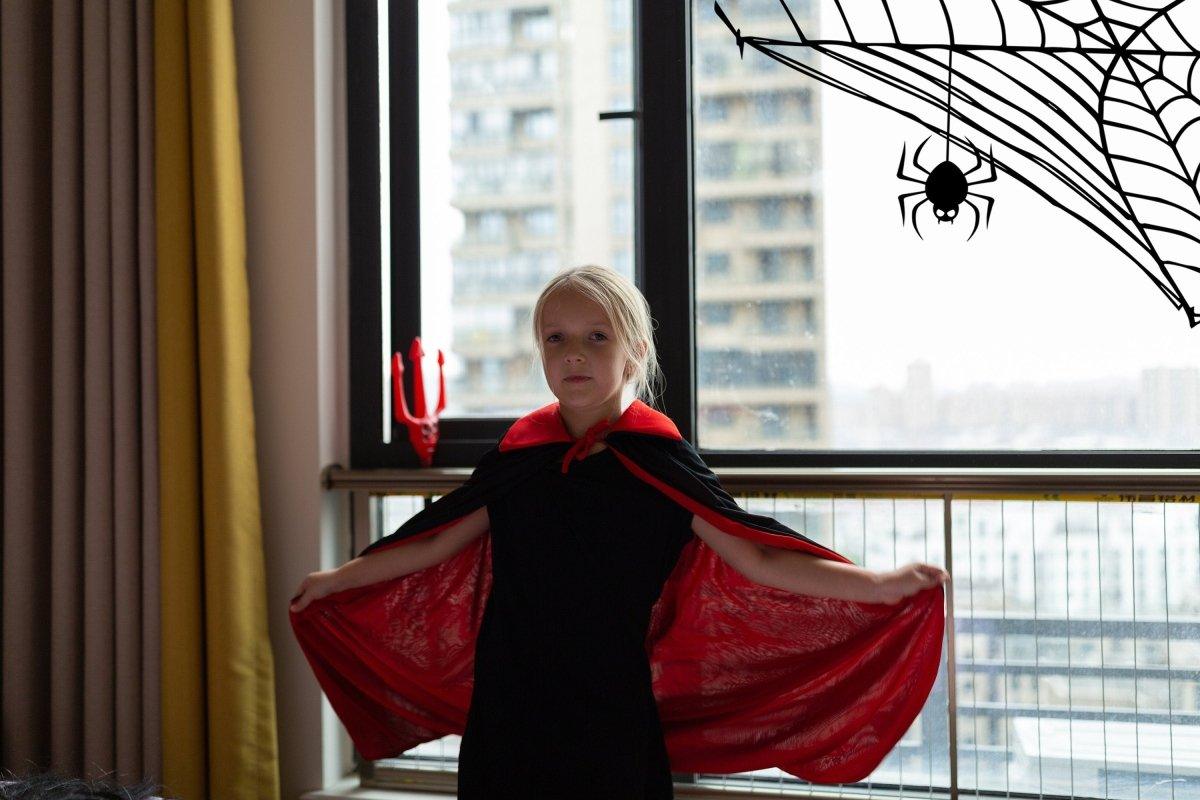 Child in a black costume with a red cape, posing in front of a Halloween spider web decal.