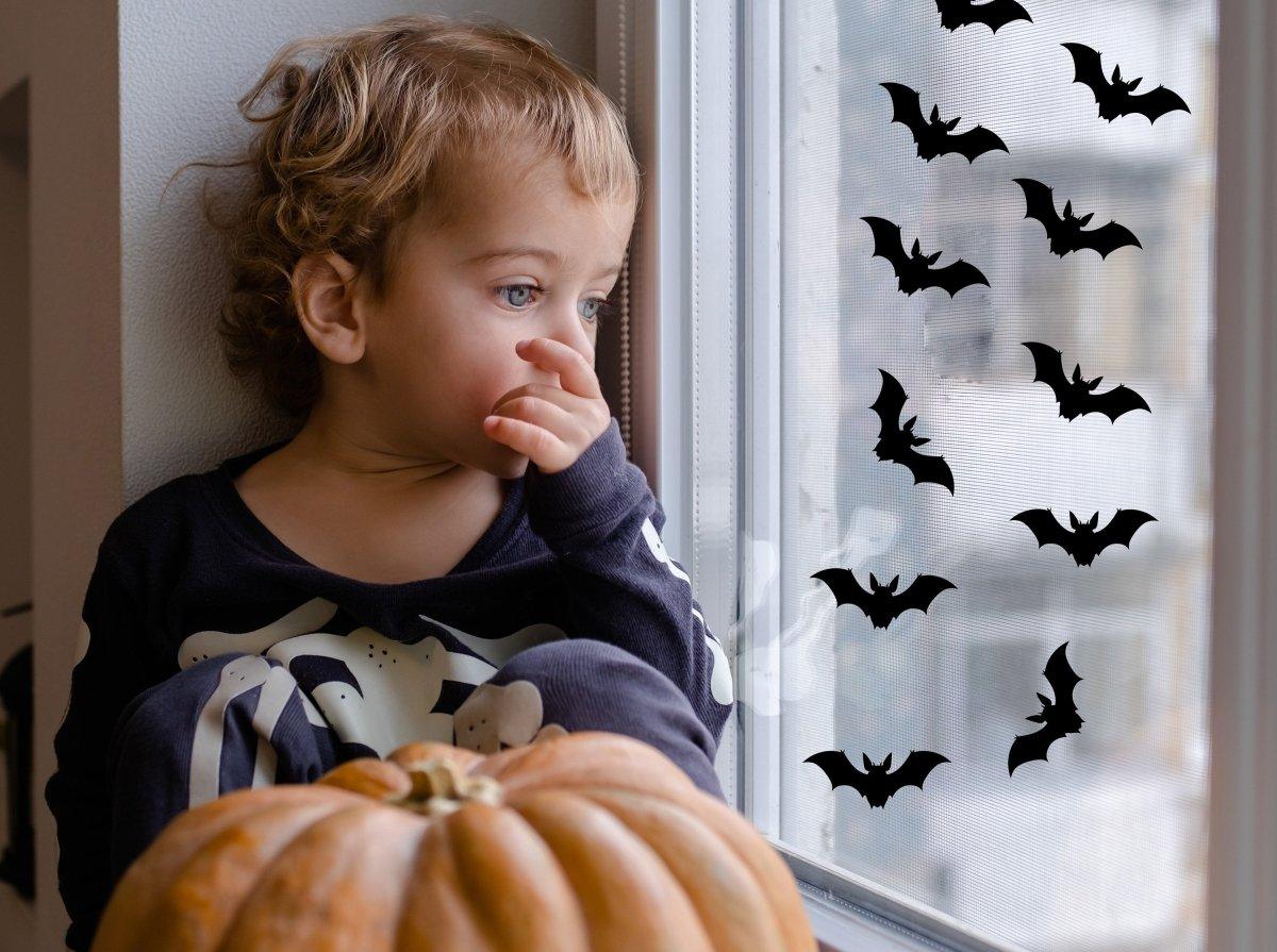 Child looking out window decorated with Halloween bat stickers, with a pumpkin on the sill.