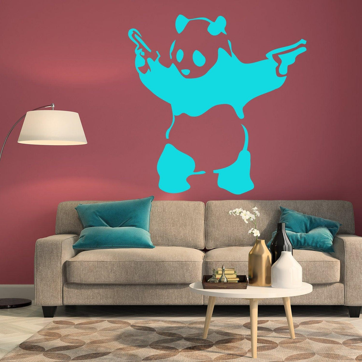 Banksy panda wall sticker in blue holding guns on pink wall above beige couch with teal cushions.