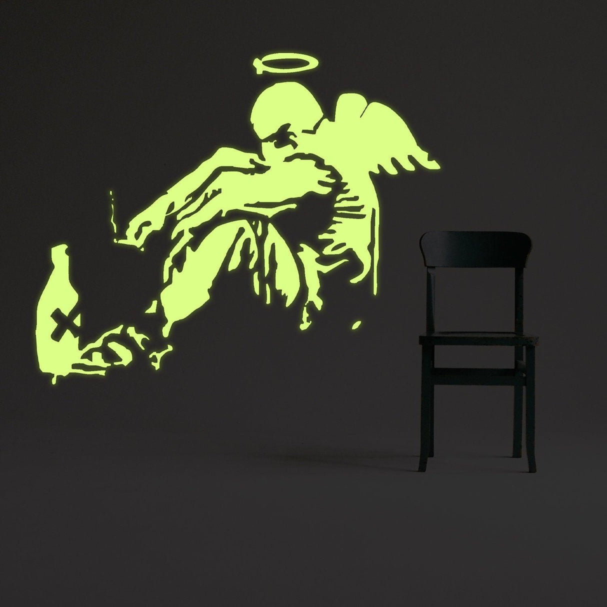 Glow in the dark Banksy angel wall sticker illuminating a dark room beside a black chair.
