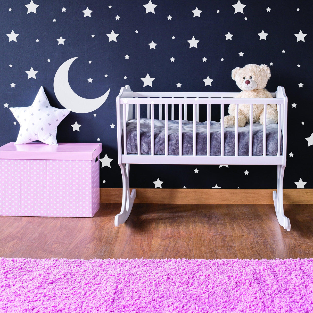 Nursery room with white stars wall decals, a crib, plush teddy bear, pink star pillow, and pink storage box.
