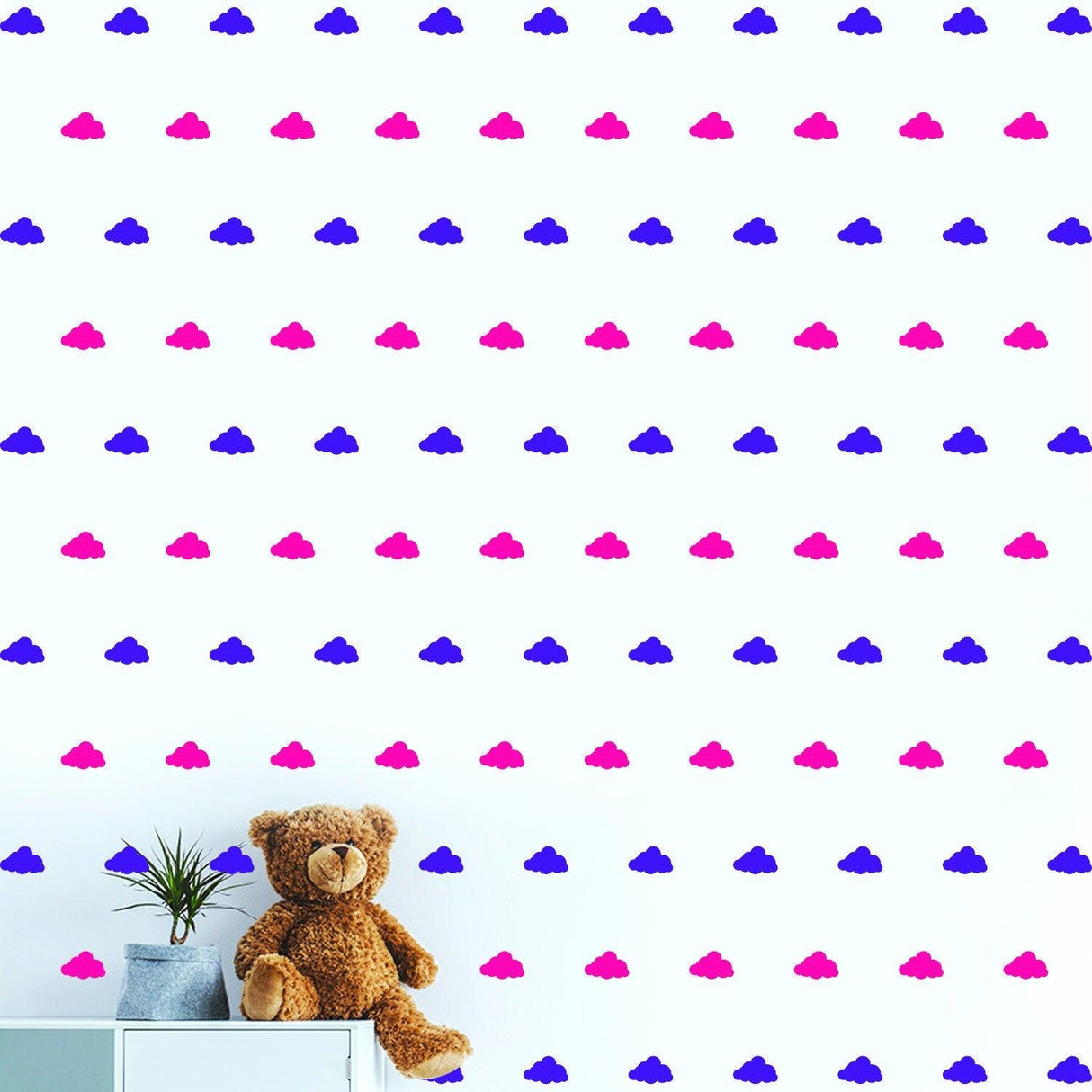 Colorful cloud wall decals in pink and blue adorn a nursery wall with a teddy bear and plant on a shelf.