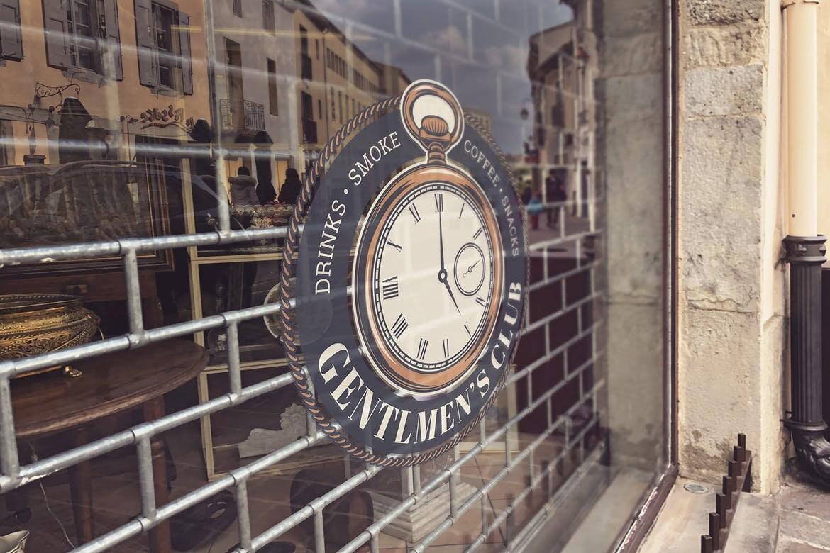 Vintage clock sign for a Gentlemen's Club, showcasing drinks, smoke, and coffee details on a shop window.