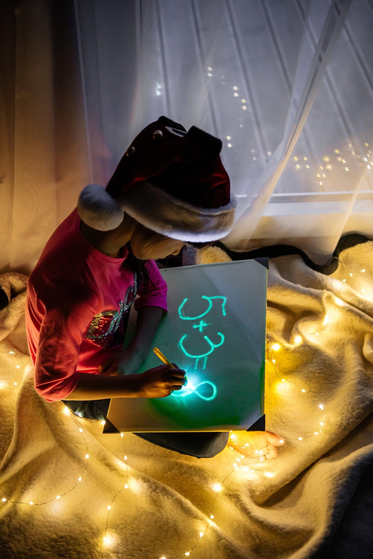 Child in a Santa hat drawing on a glowing board surrounded by fairy lights, showcasing creativity and fun.