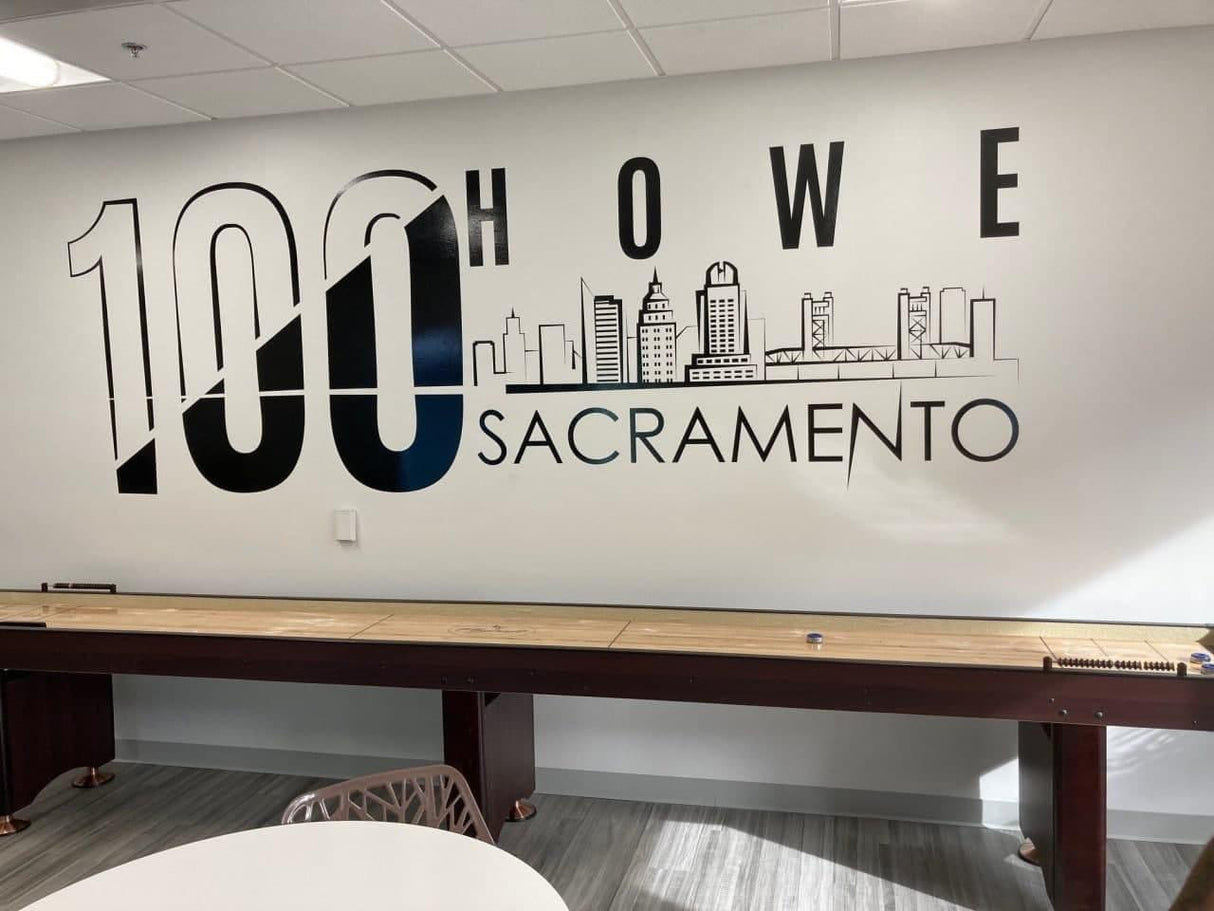 Wall mural featuring '100 Howe Sacramento' with skyline design and wooden table in a modern office space.