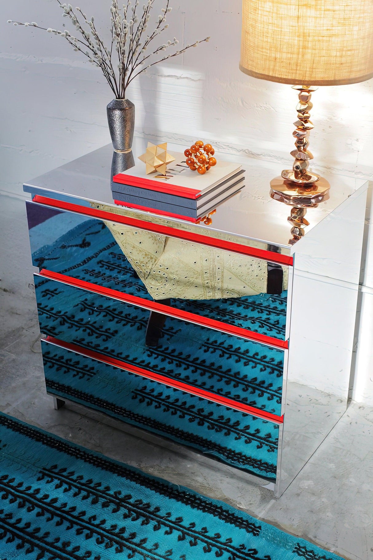 Stylish mirrored metallic silver chrome dresser with vibrant decor, lamp, and patterned rug in modern interior.