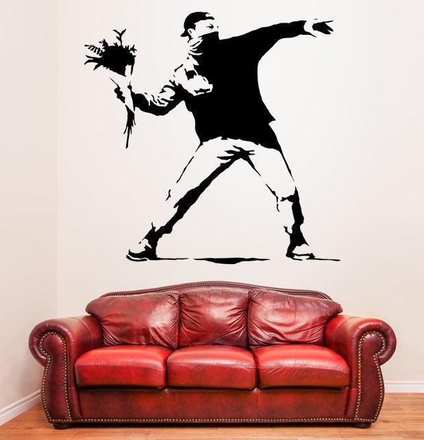 Banksy Flower Thrower wall decal on a white wall above a red leather couch, showcasing urban art and graffiti decor.