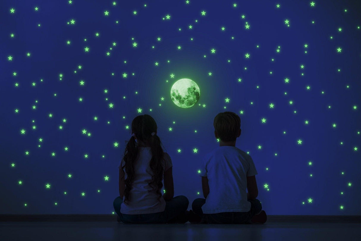 Children sitting on the floor admiring glowing stars and moon decals on a dark wall in a nursery.
