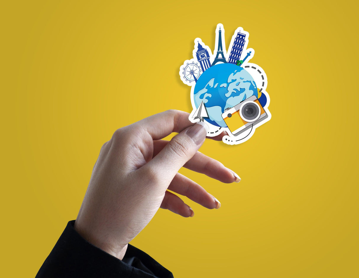 Hand holding a colorful travel themed die cut sticker featuring landmarks and a camera against a yellow background.
