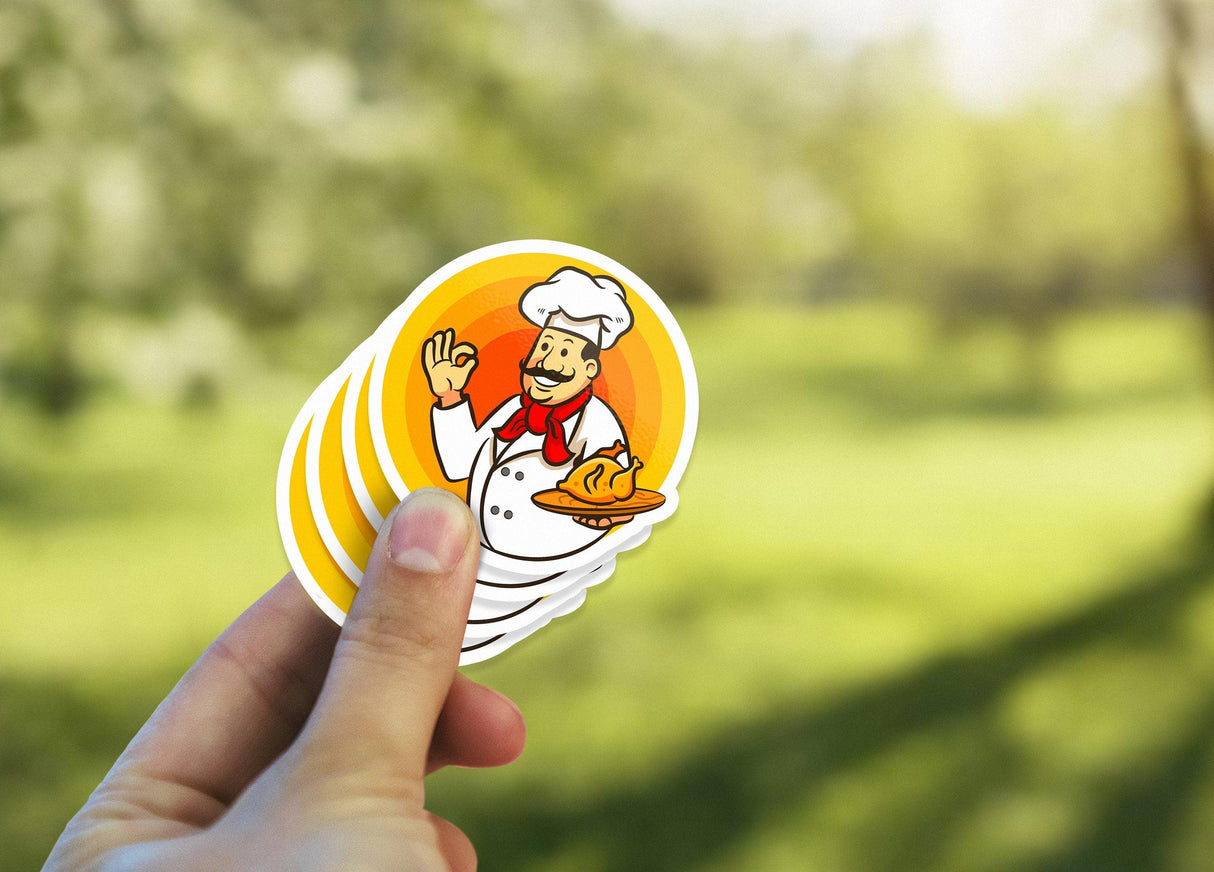 Hand holding colorful laminated kiss die cut sticker featuring a cheerful chef with a pastry.