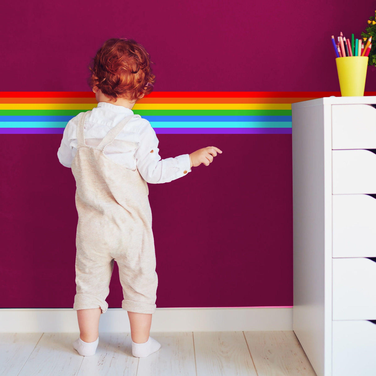 Child pointing at colorful rainbow border sticker on purple wall, enhancing room decor with playful design.