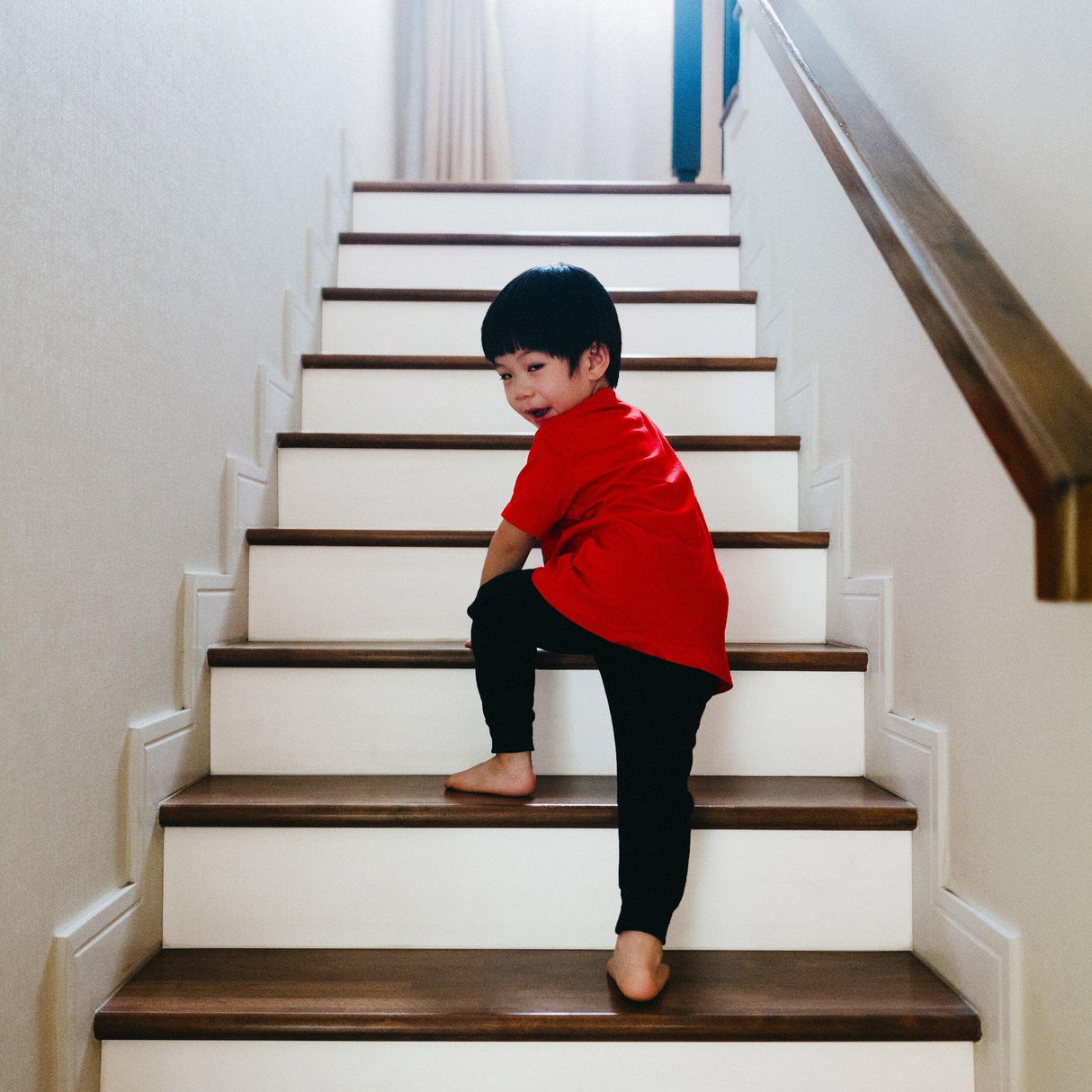 Young child in a red shirt playfully climbing white stairs with wooden risers.
