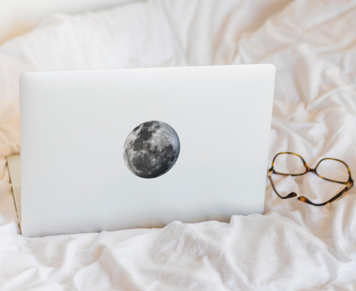 Glow in the dark laptop decal featuring a full moon design on a white laptop, resting on a cozy blanket.