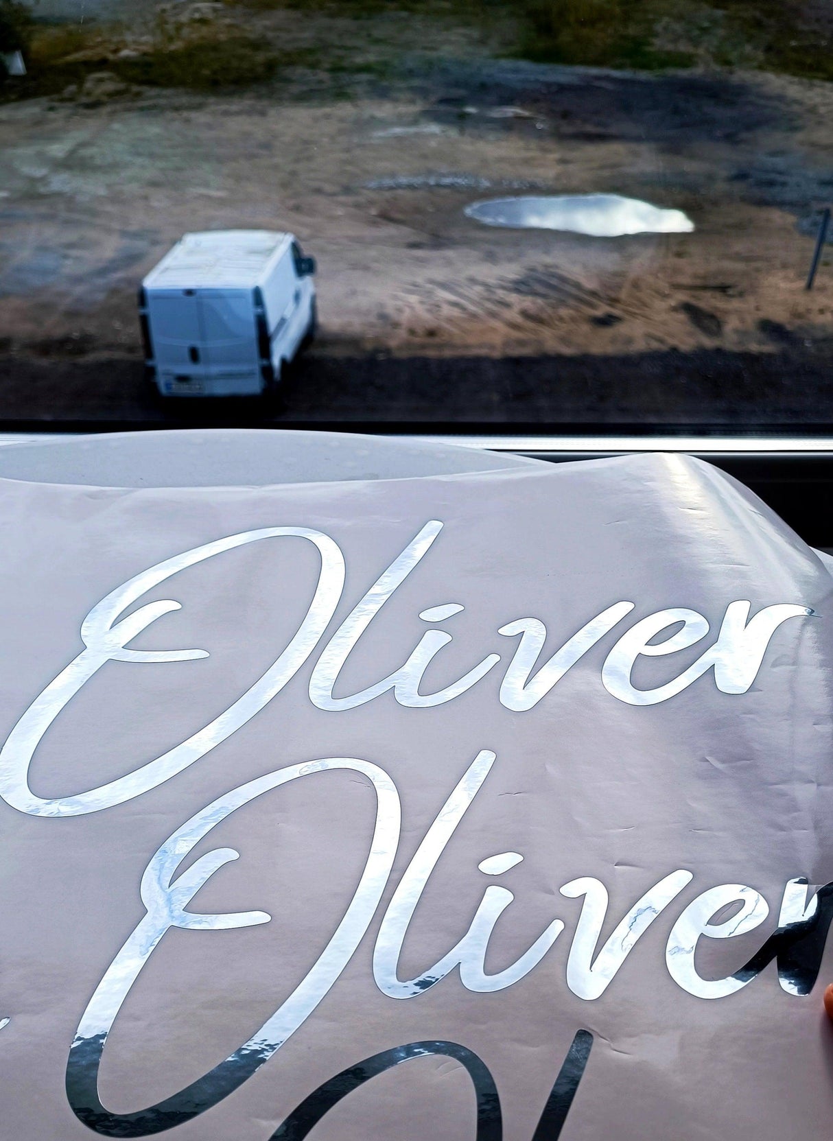 Personalized vinyl name sticker in silver chrome finish with the name 'Oliver' on a backdrop of an outdoor scene.