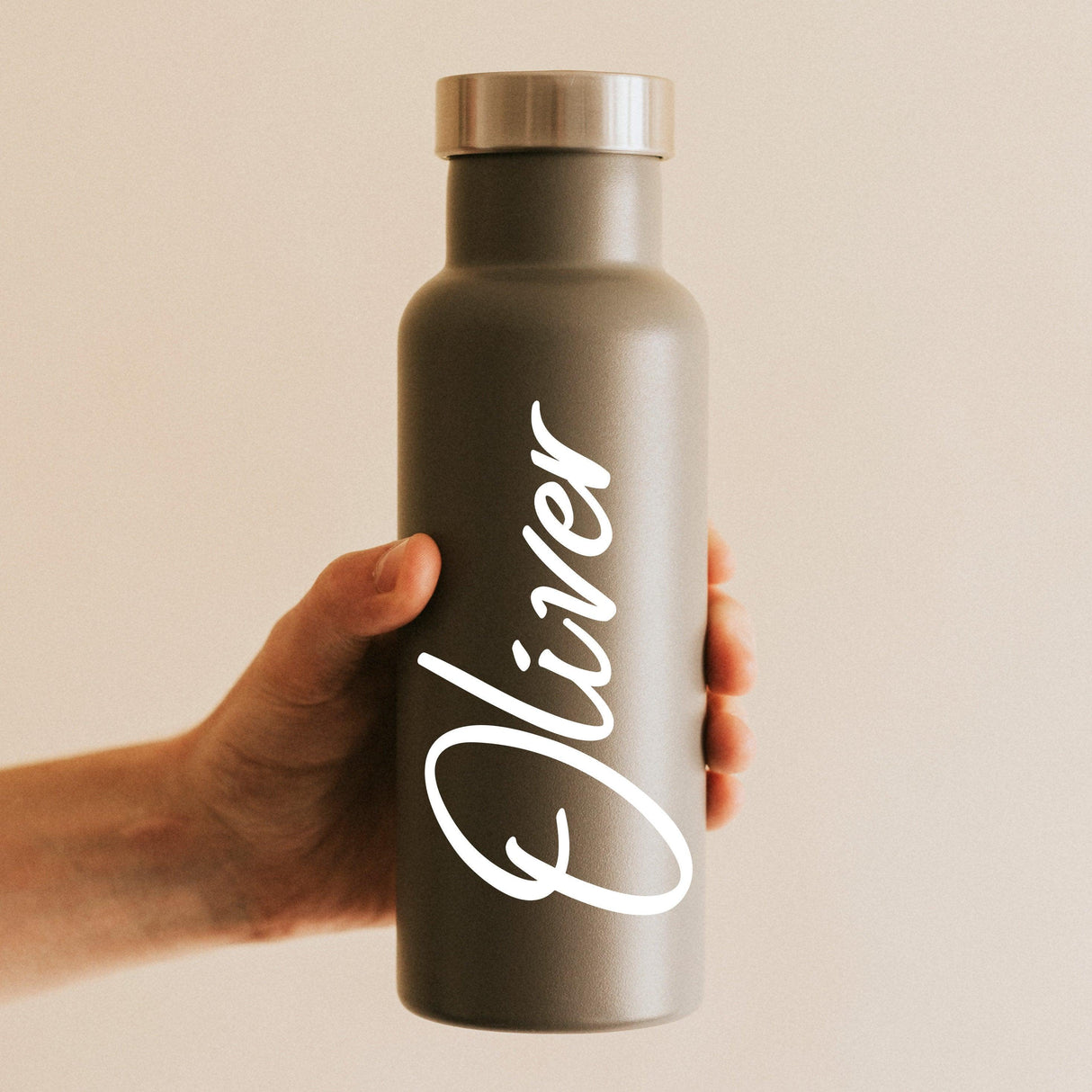 Personalized vinyl name decal on a gray water bottle being held, featuring the name 'Oliver' in stylish lettering.