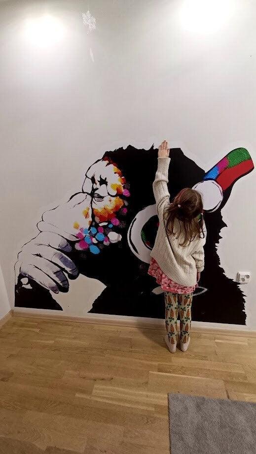 Child reaching toward a colorful Banksy-inspired monkey mural sticker on a white wall.