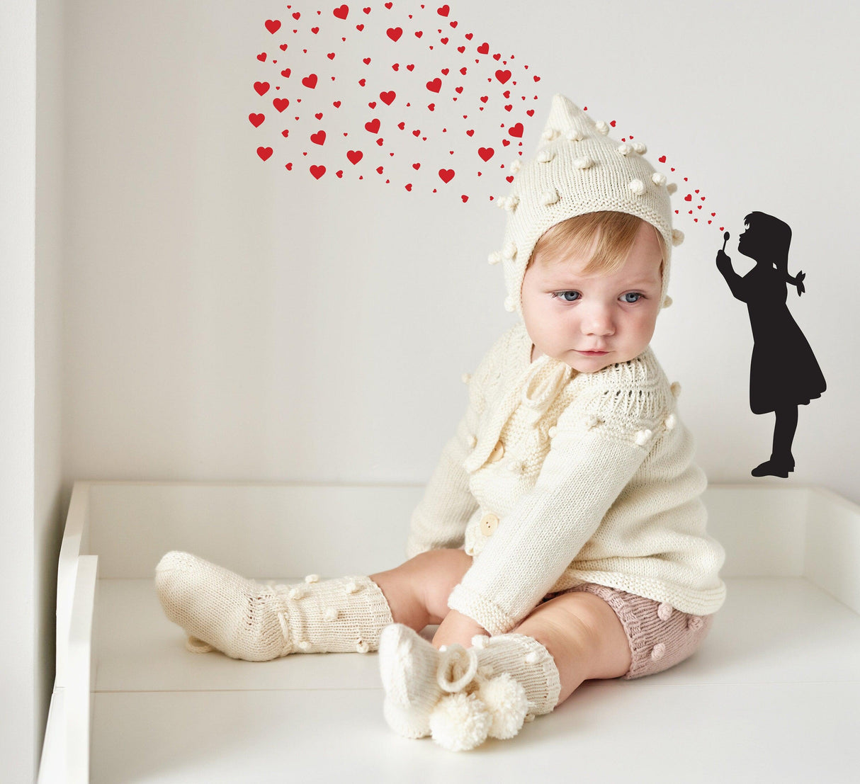 Cute baby girl in knitted outfit with heart bubbles and graffiti style girl silhouette on wall.
