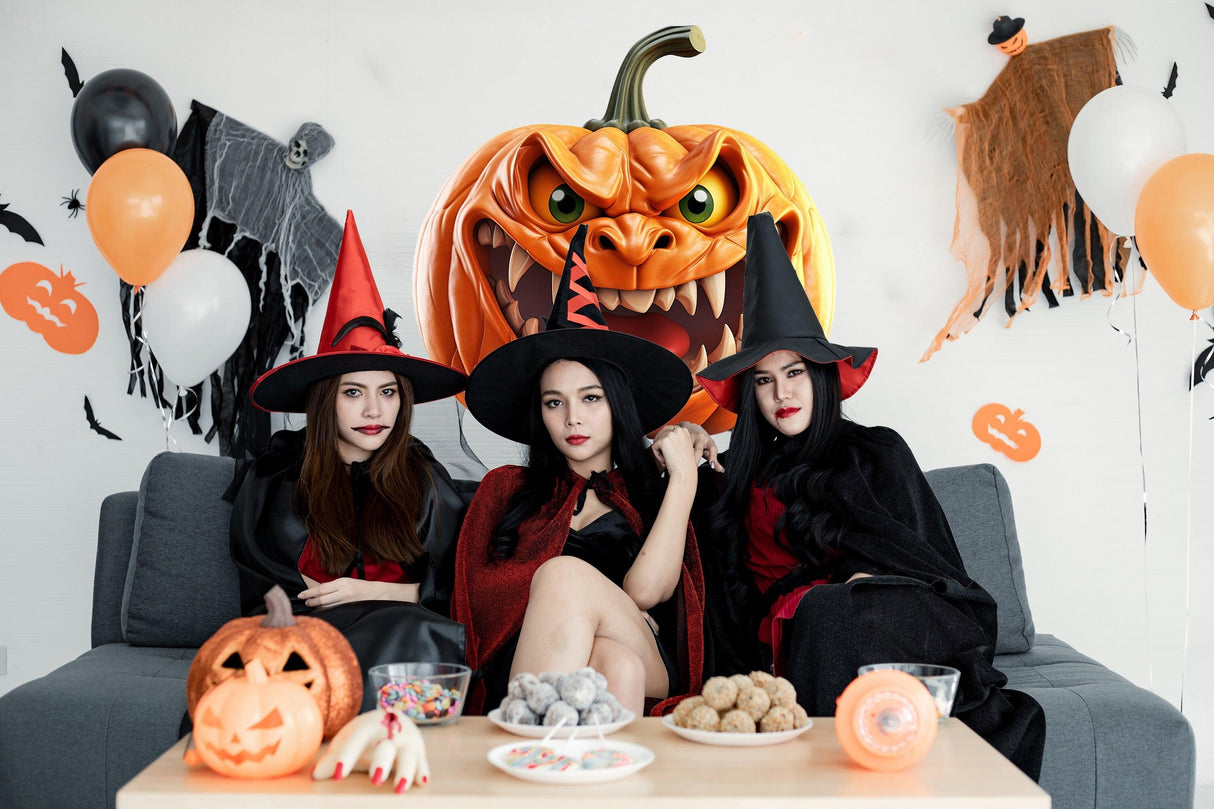 Three women in witch costumes pose with Halloween decorations, including a large pumpkin and festive treats.