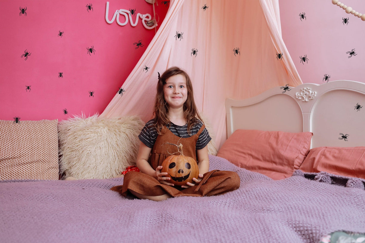Girl sitting on a bed with spider wall decals, holding a Halloween pumpkin, in a cozy decorated room.
