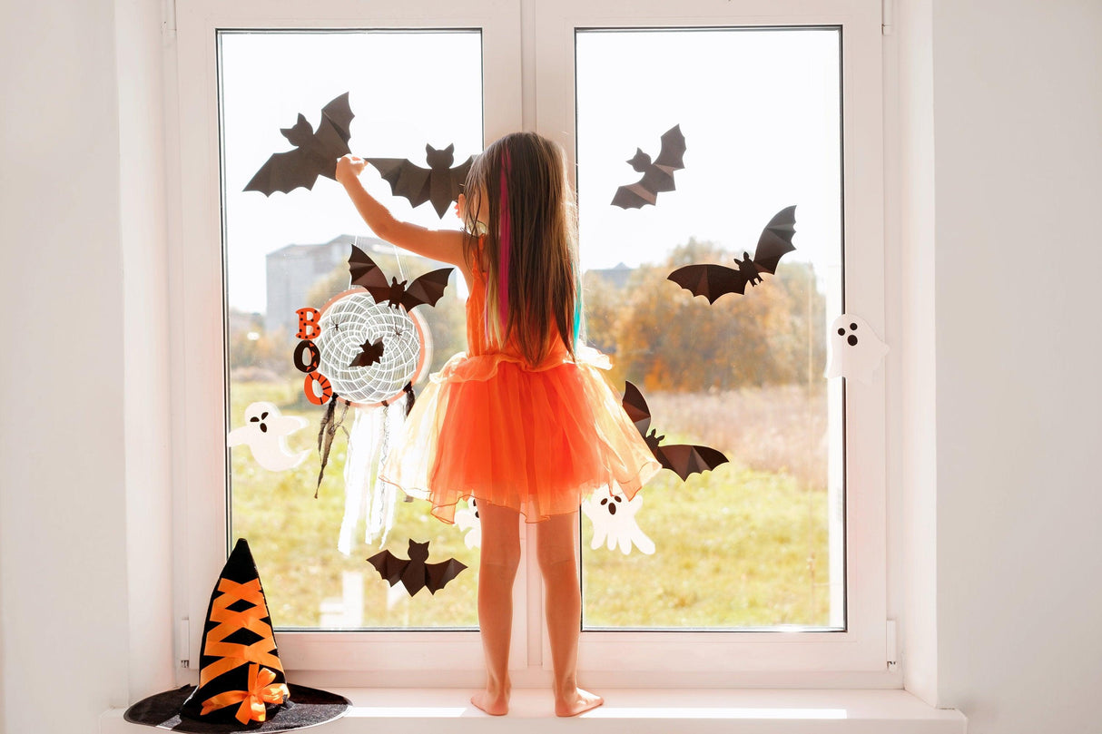 Child in an orange dress decorating window with Halloween bat stickers, creating a spooky atmosphere.