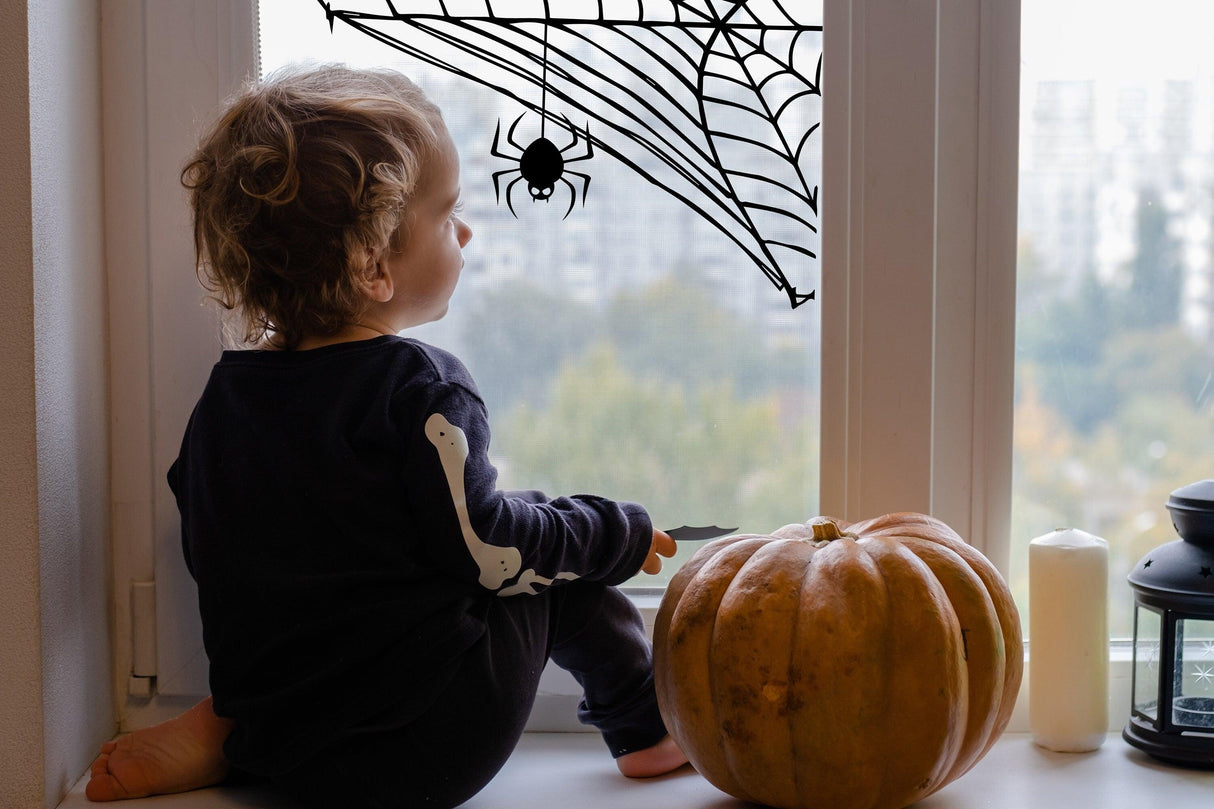 Child sitting by window with Halloween spider web decal and a pumpkin, creating a spooky atmosphere.