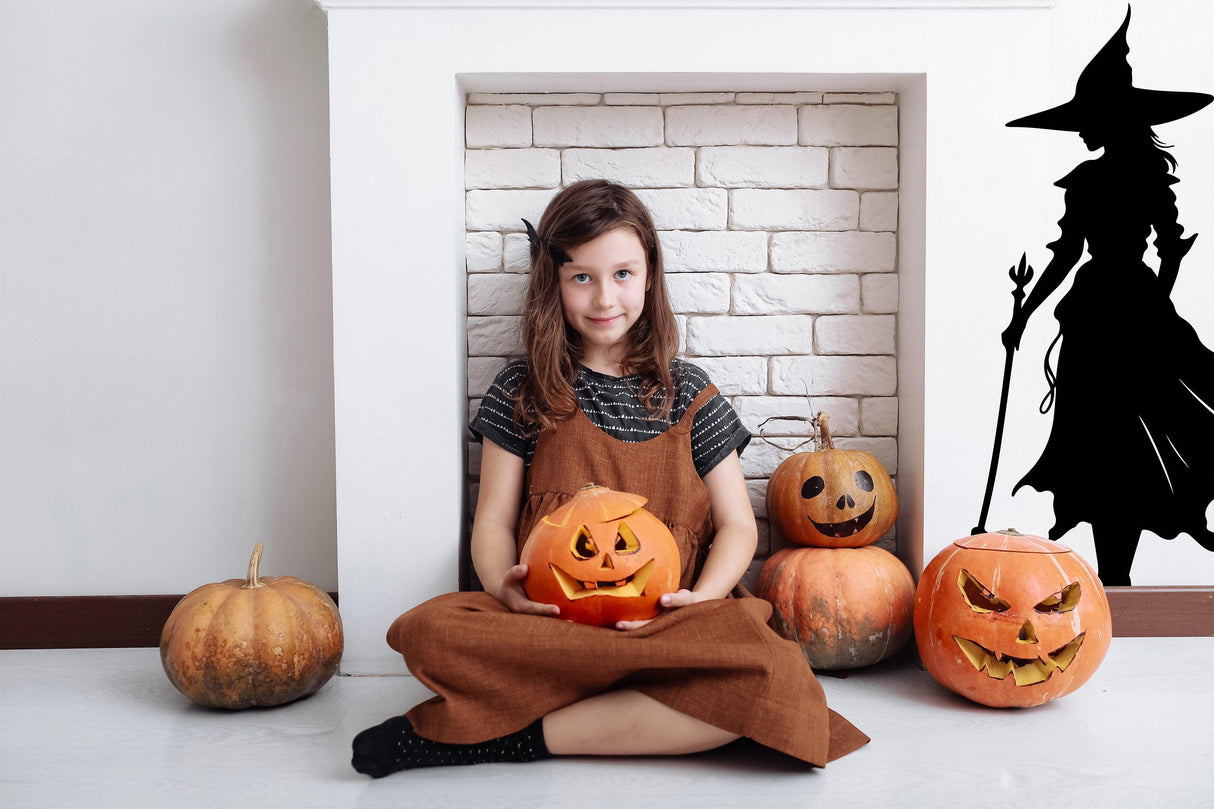Girl sitting with carved pumpkins and a witch silhouette decal, celebrating Halloween.
