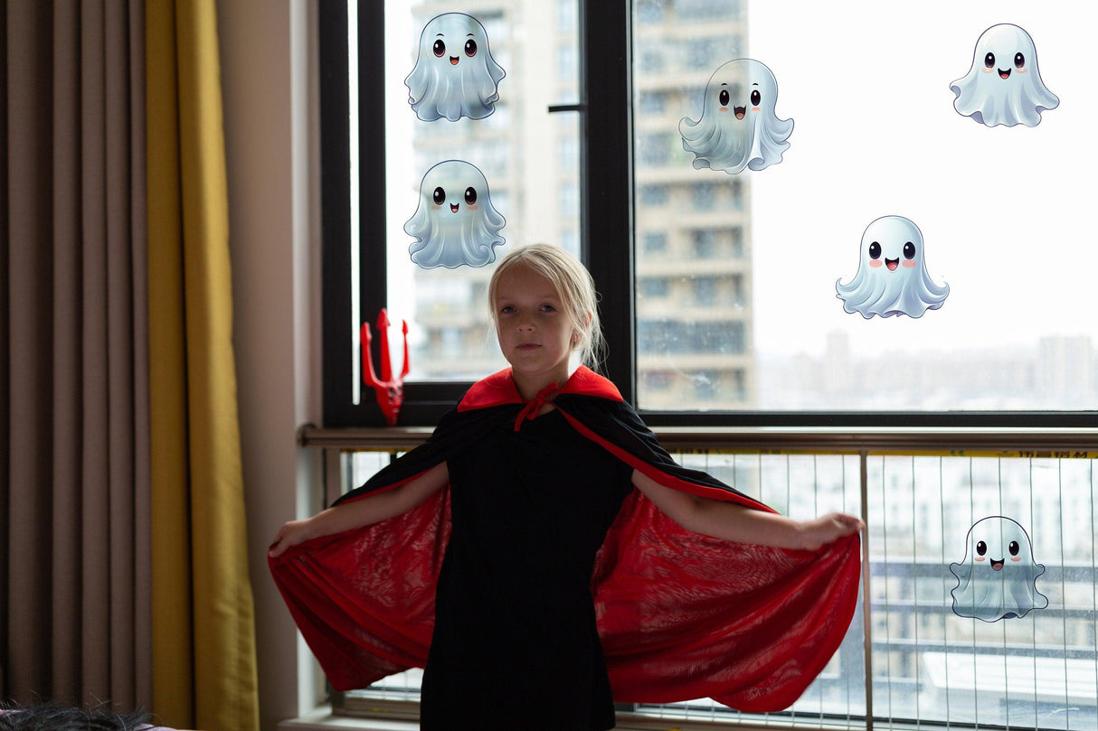 Child in a black costume with a red cape standing by a window decorated with frosted ghost stickers for Halloween.