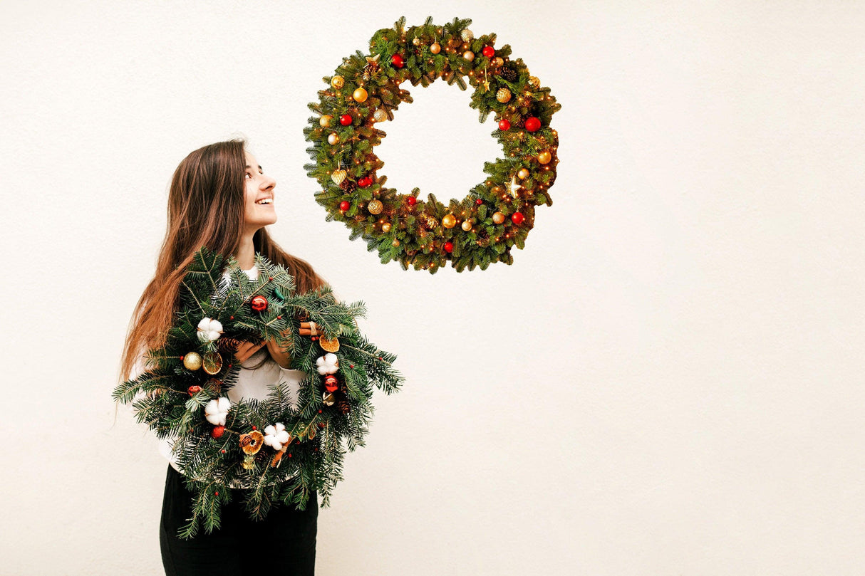 Woman holding a Christmas wreath while smiling at another hanging wreath on the wall, festive holiday decor.