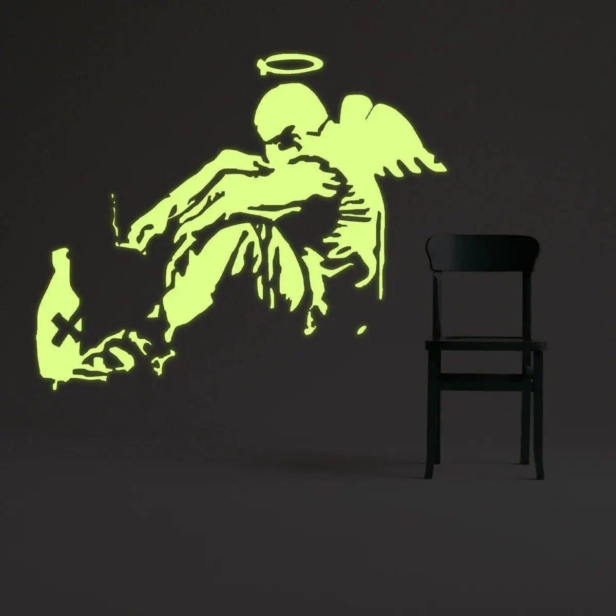 Glowing Banksy fallen angel wall sticker decal illuminating a dark room beside a black chair.