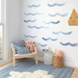Watercolor wave wall decals in varying shades of blue arranged on a white wall above a cozy chair and storage baskets in a coastal-themed kids&#39; room.