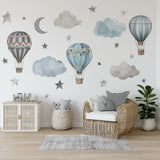 A boys’ nursery decorated with watercolor-style wall decals featuring three hot air balloons in muted blue and beige tones, surrounded by fluffy clouds, gray stars, and a crescent moon above a rattan chair and soft rug.