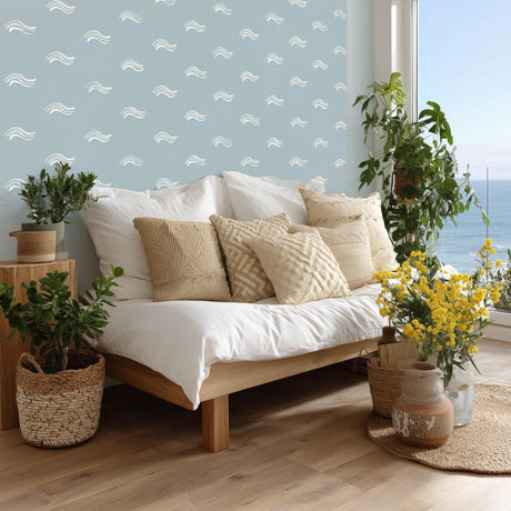 A serene coastal bedroom with white ocean wave decals scattered across a soft blue wall. The decor includes natural wood furniture, neutral boho pillows, potted plants, and a view of the ocean through a large window, creating a relaxed beach cottage feel.